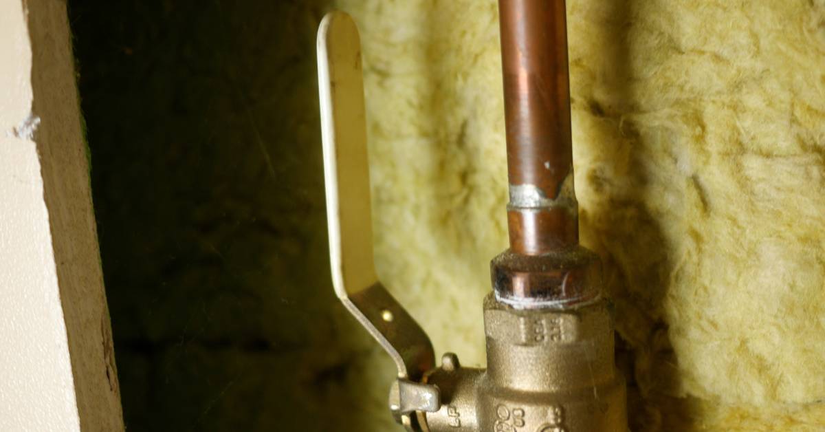 A copper-colored pipe connecting to a brass lever valve between a piece of drywall and yellow insulation.