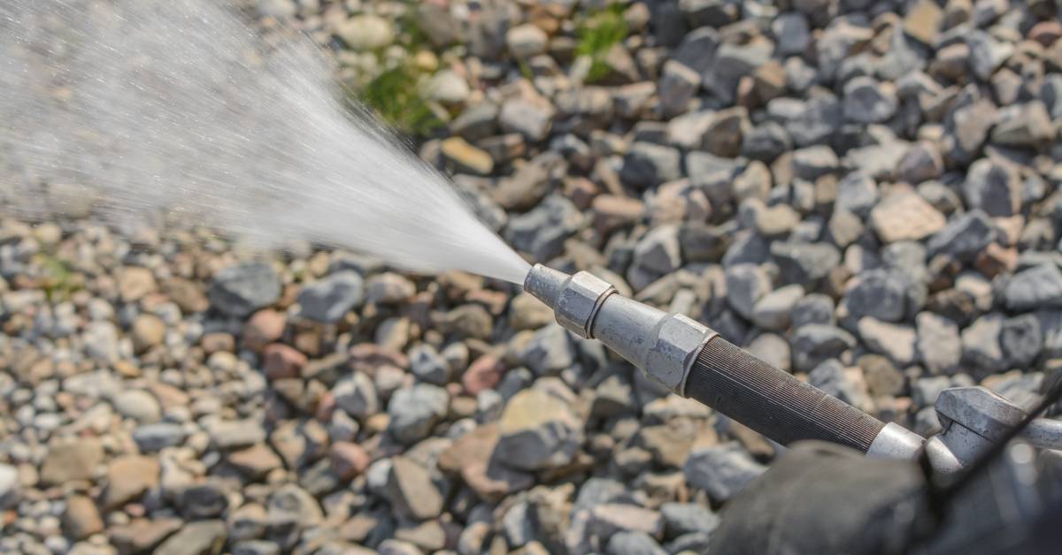 A close-up of the end of a fire hose spraying high-pressure water across a background of small rocks.